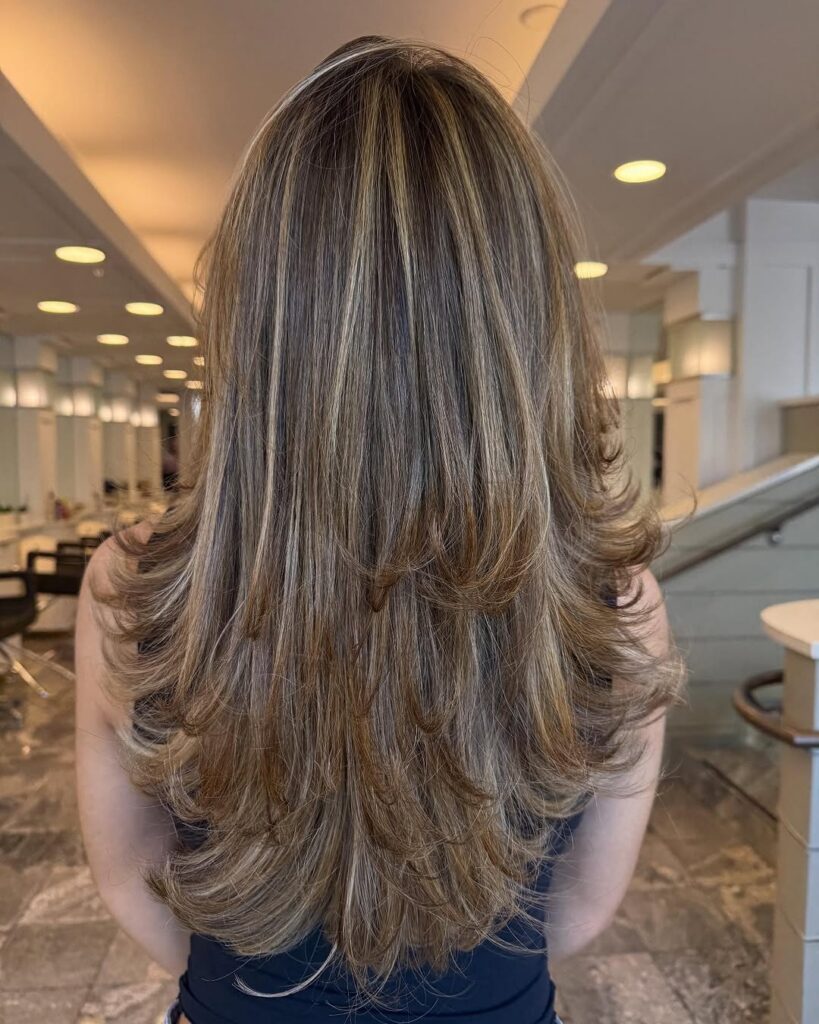 Back view of a woman with long, layered, highlighted brown hair in a salon setting.