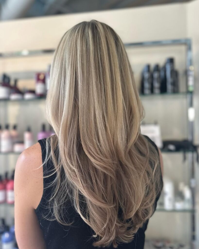 Back view of a person with long layered blonde hair cascading down the back in a salon setting with shelves of hair products behind them.